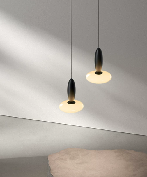 Two modern pendant lights with black teardrop-shaped centers and glowing warm white circular shades, hanging against a gray wall above a bed.