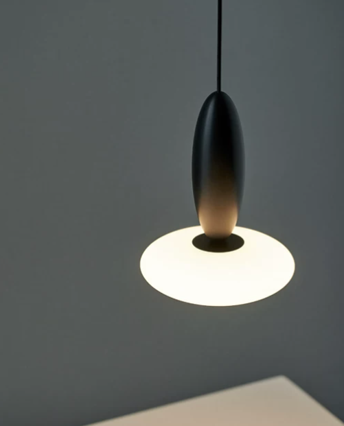 A modern pendant light with a matte black elongated shade and a glowing white circular diffuser, suspended against a dark gray wall.